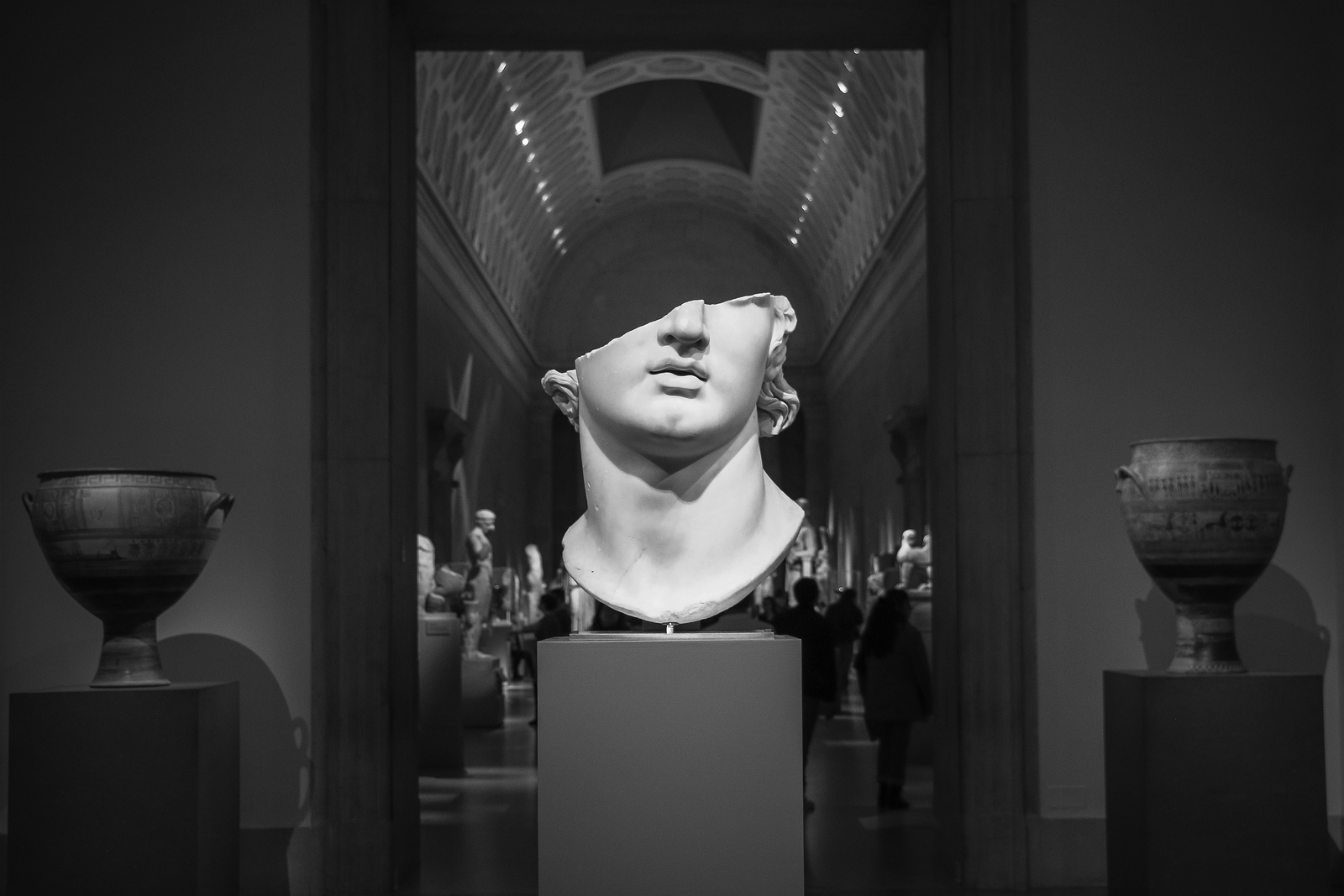 Black and white photo of a large broken marble head sculpture in a museum gallery, with vases on either side.
